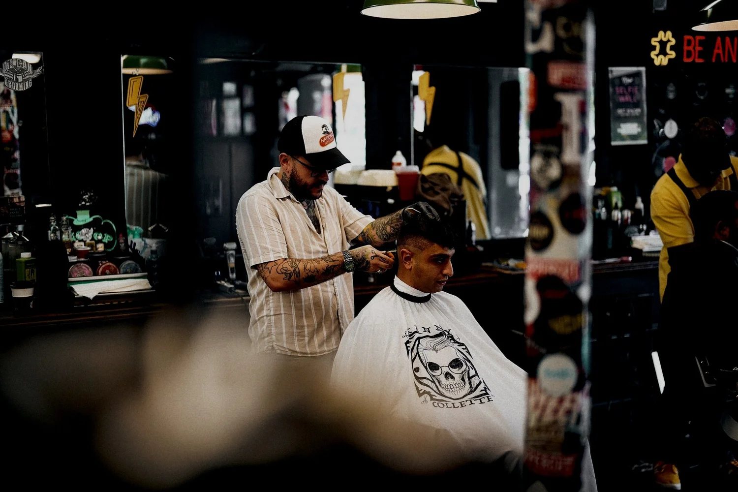 A barber in a cap working on a client’s fade in the middle of the shop.