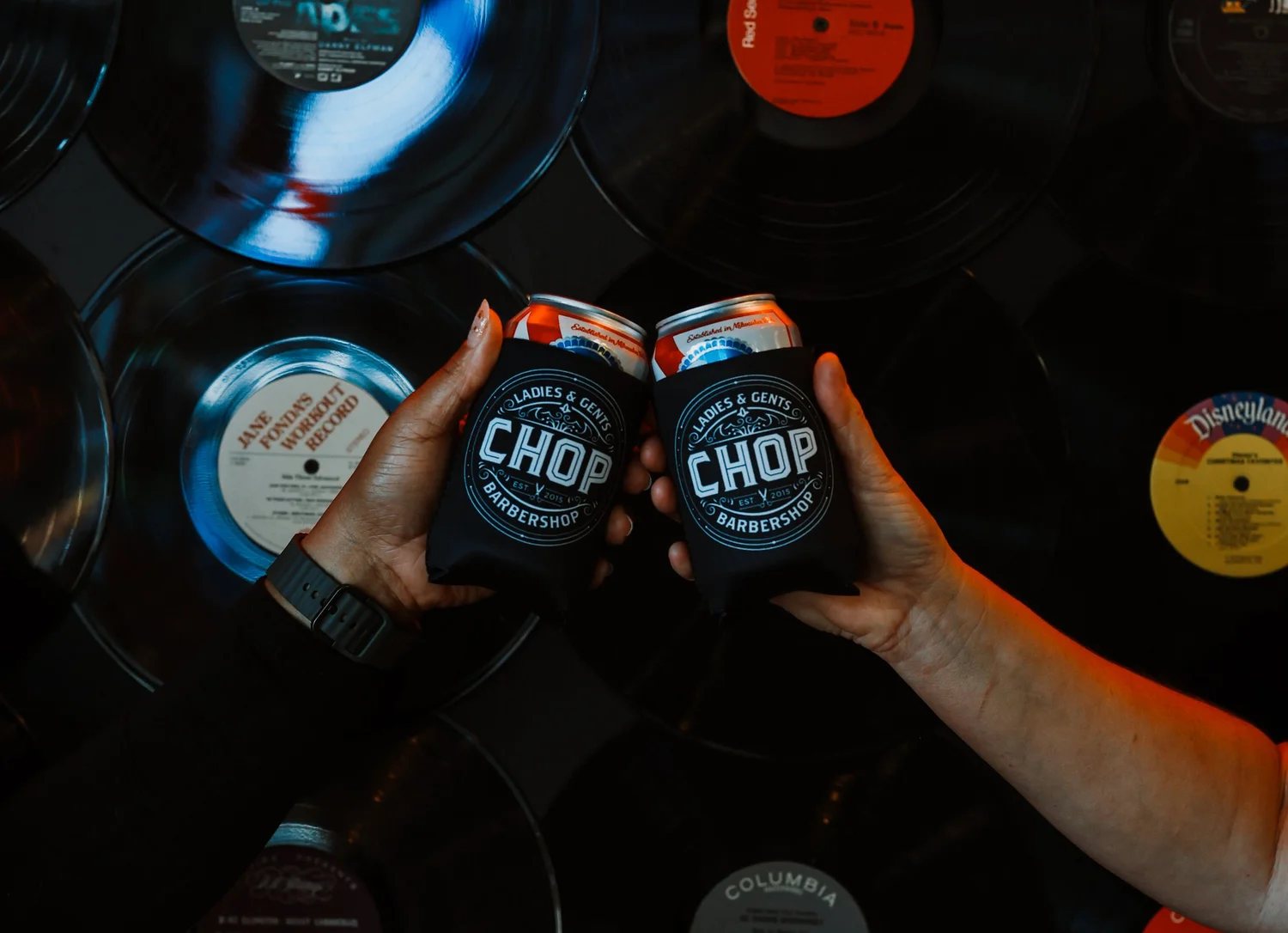 Two hands touching CHOP can koozies together above a wall of vinyl records.